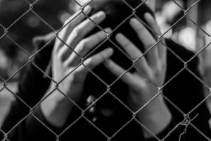 juvenile behind a chain link fence with head in their hands. Black and white.
