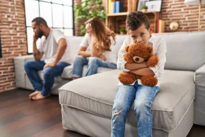 Parents fighting on couch behind a son hugging a teddy bear.
