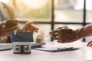 A miniature house on the table with two people talking over paperwork.